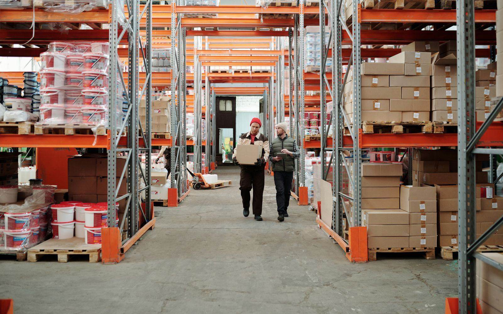 Trabajadores de almacén revisando y trasladando cajas entre estanterías, representando la logística y distribución.
